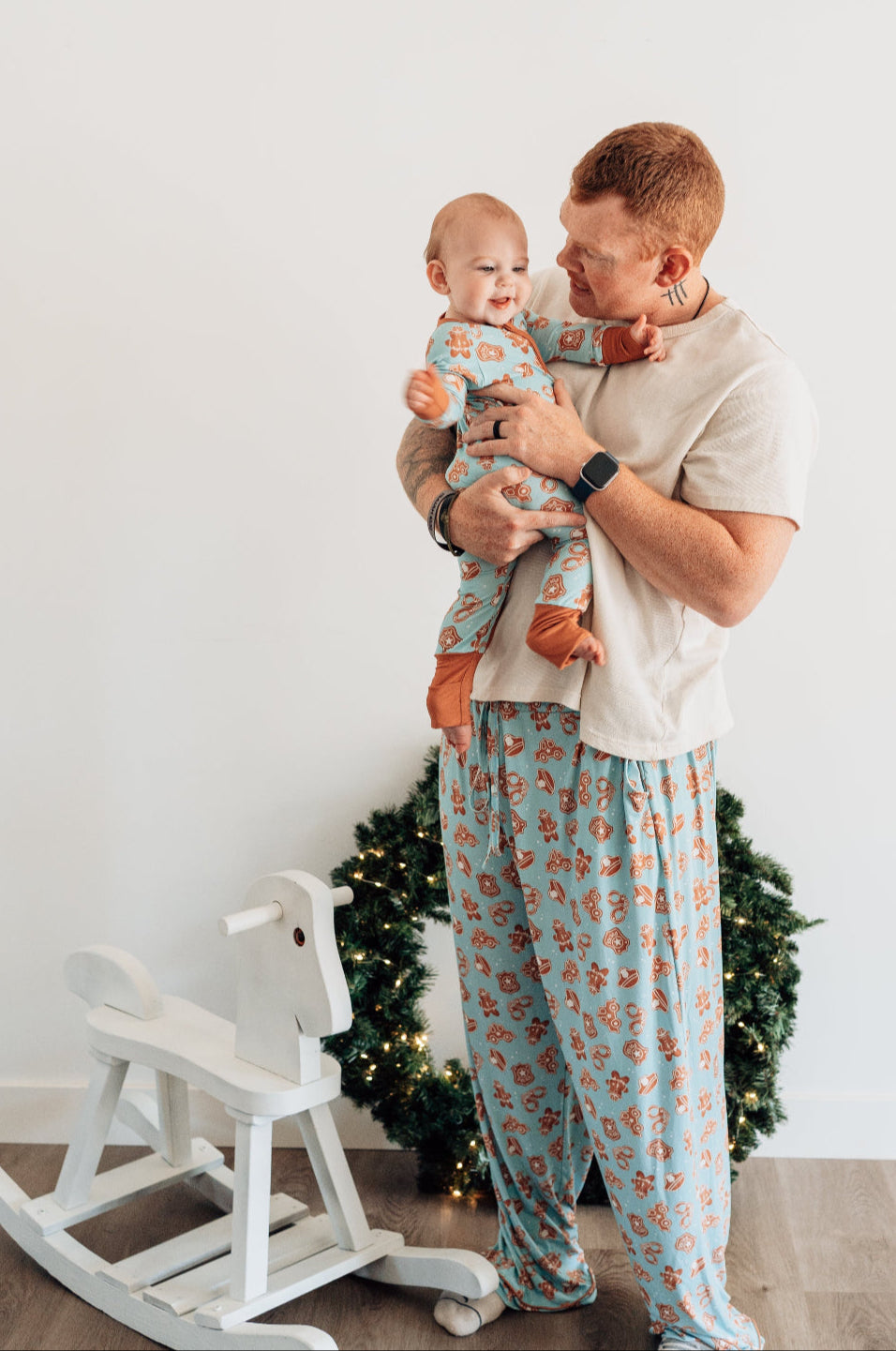 Man holding a baby in a room with a white wall, wooden floor, and a white rocking horse.