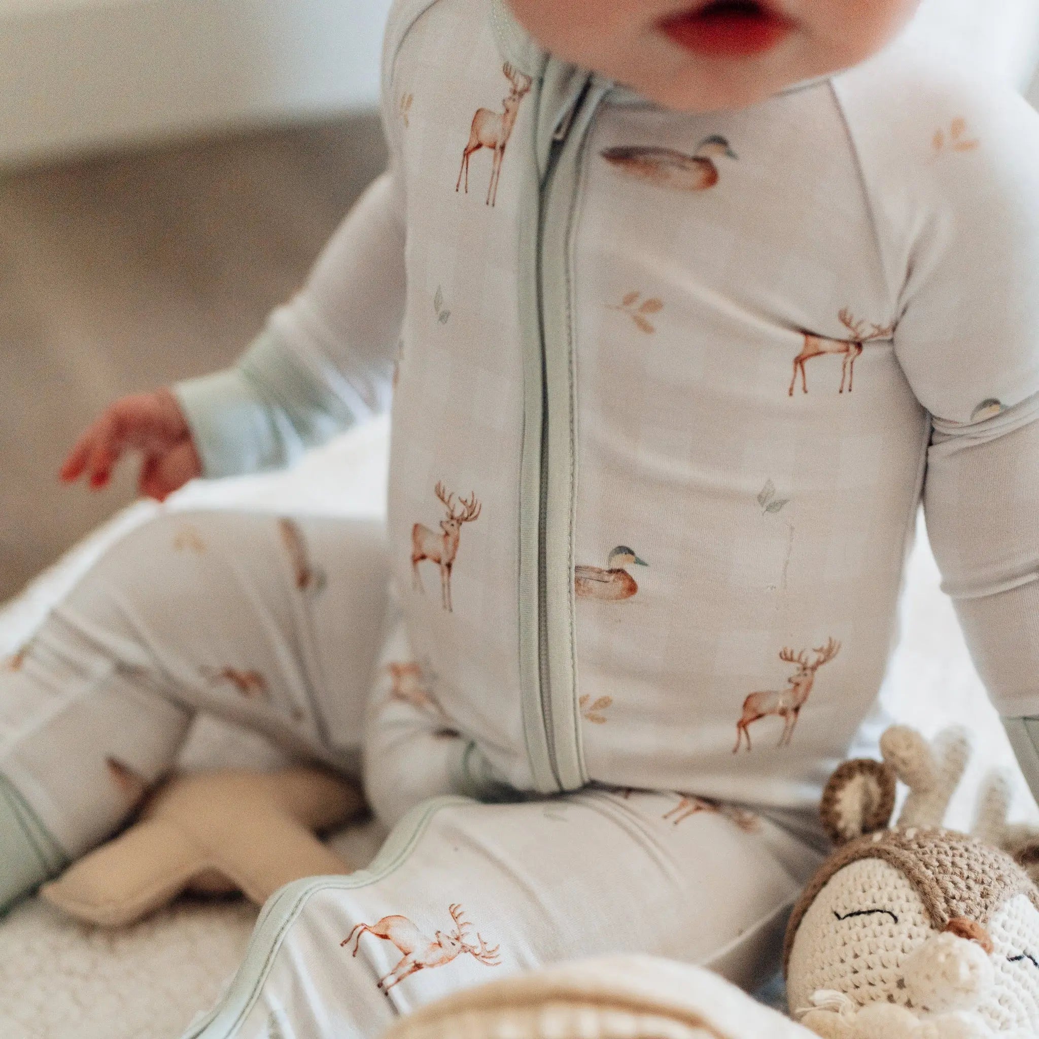 Baby in a light gray onesie with animal prints, surrounded by soft toys on a neutral background.