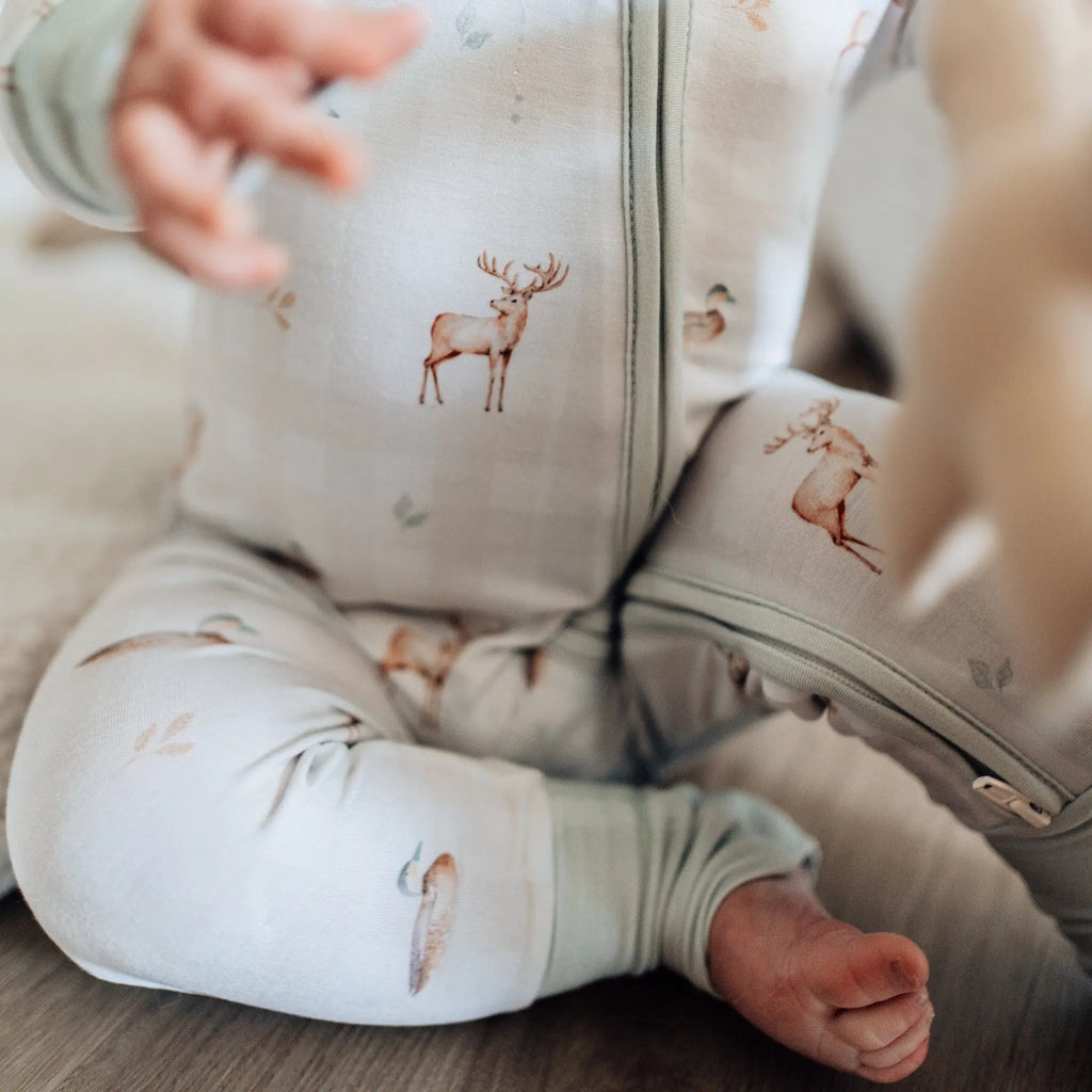 Baby in a light-colored onesie with deer patterns sitting on a wooden floor.