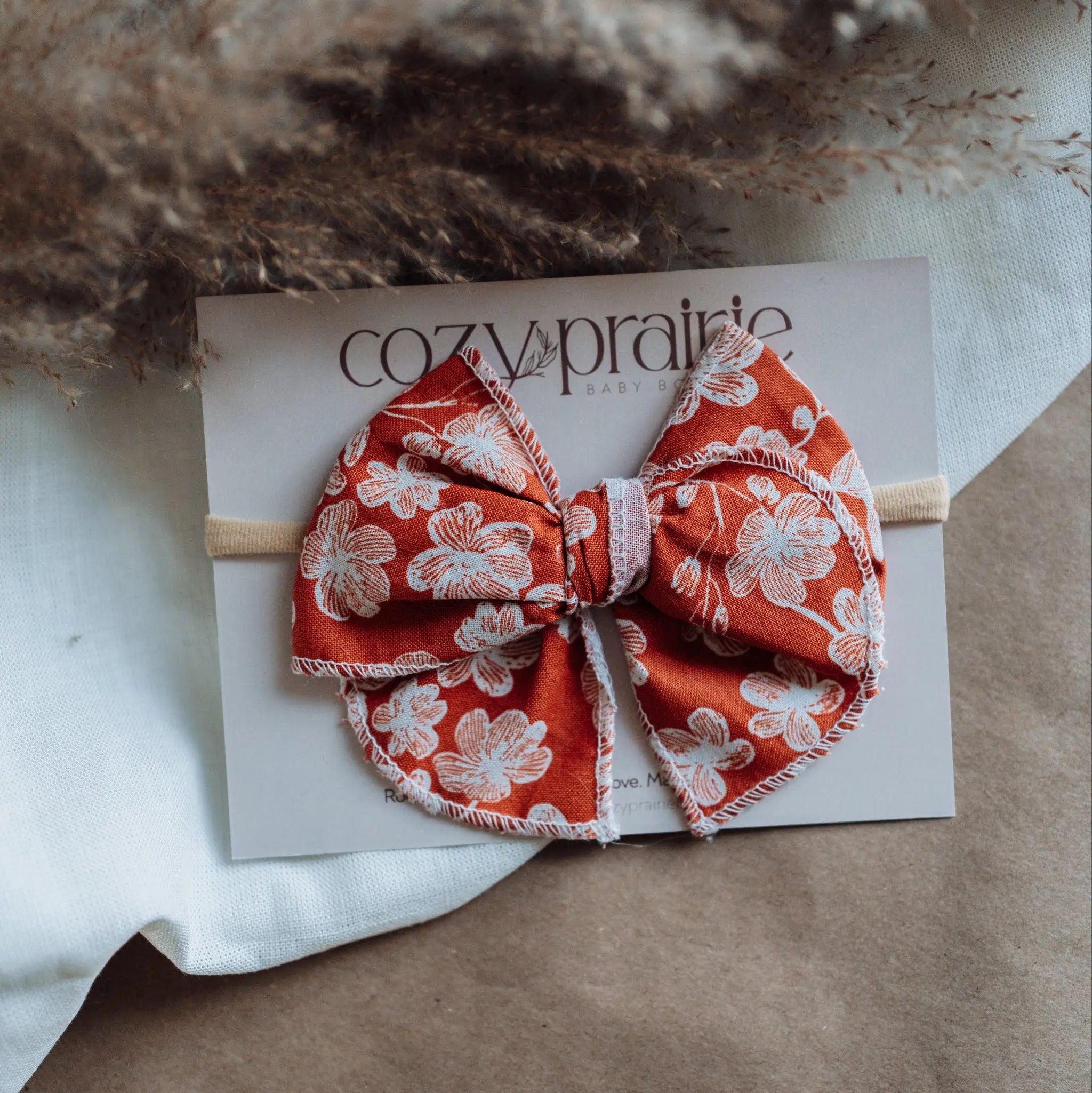 Red floral bow hair accessory on a card with 'cozy prairie' branding, placed on a textured surface.