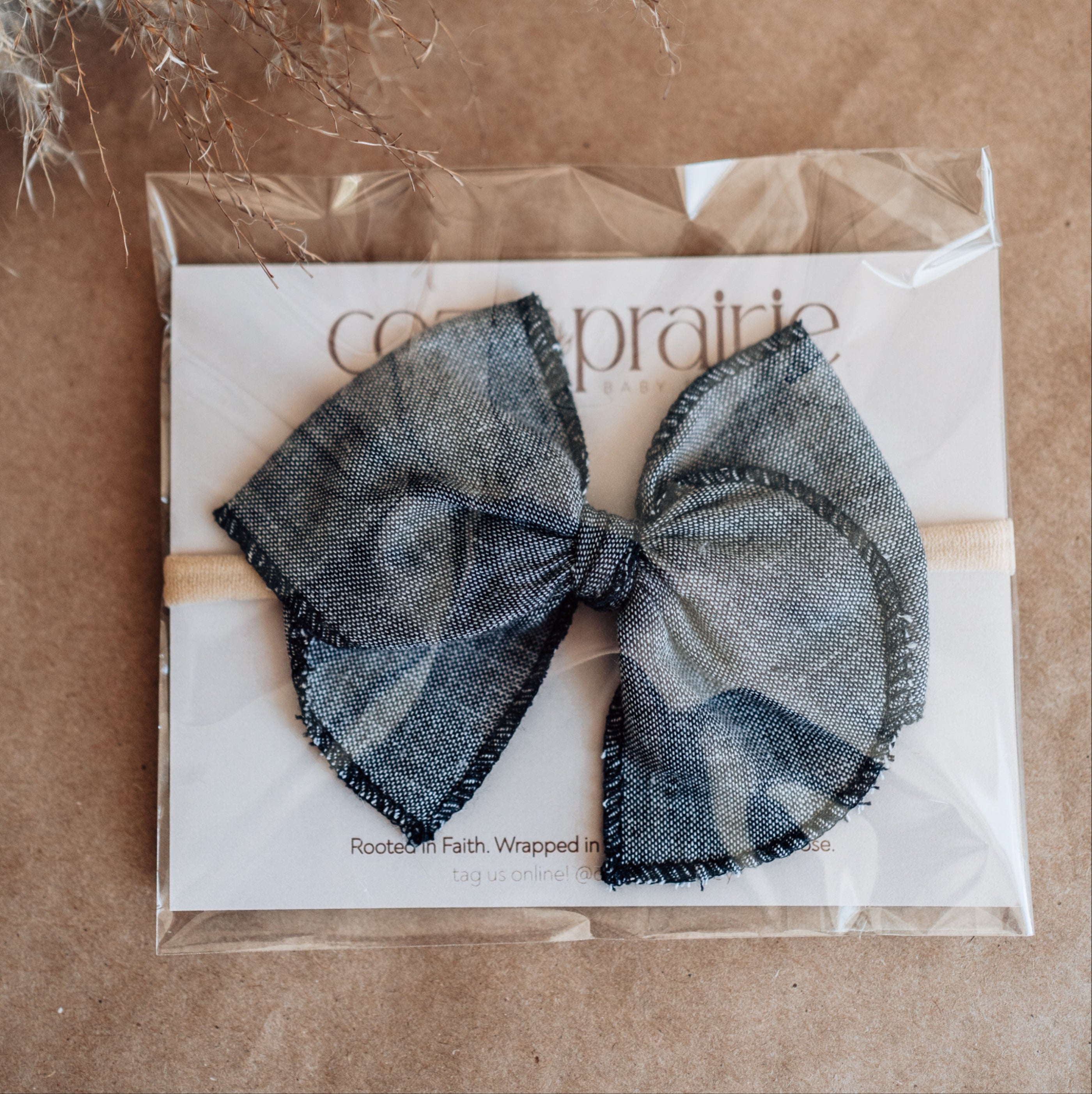 Packaged black bow hair accessory on brown paper with dried plants.