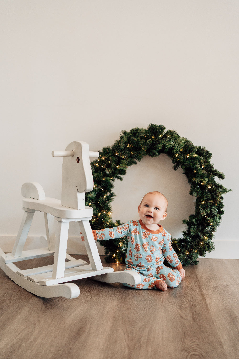 Baby sitting on a wooden floor next to a white rocking horse with a Christmas wreath in the background.