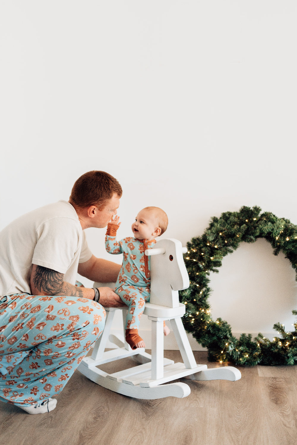 Man holding a baby in a room with a white wall, wooden floor, and a white rocking horse.