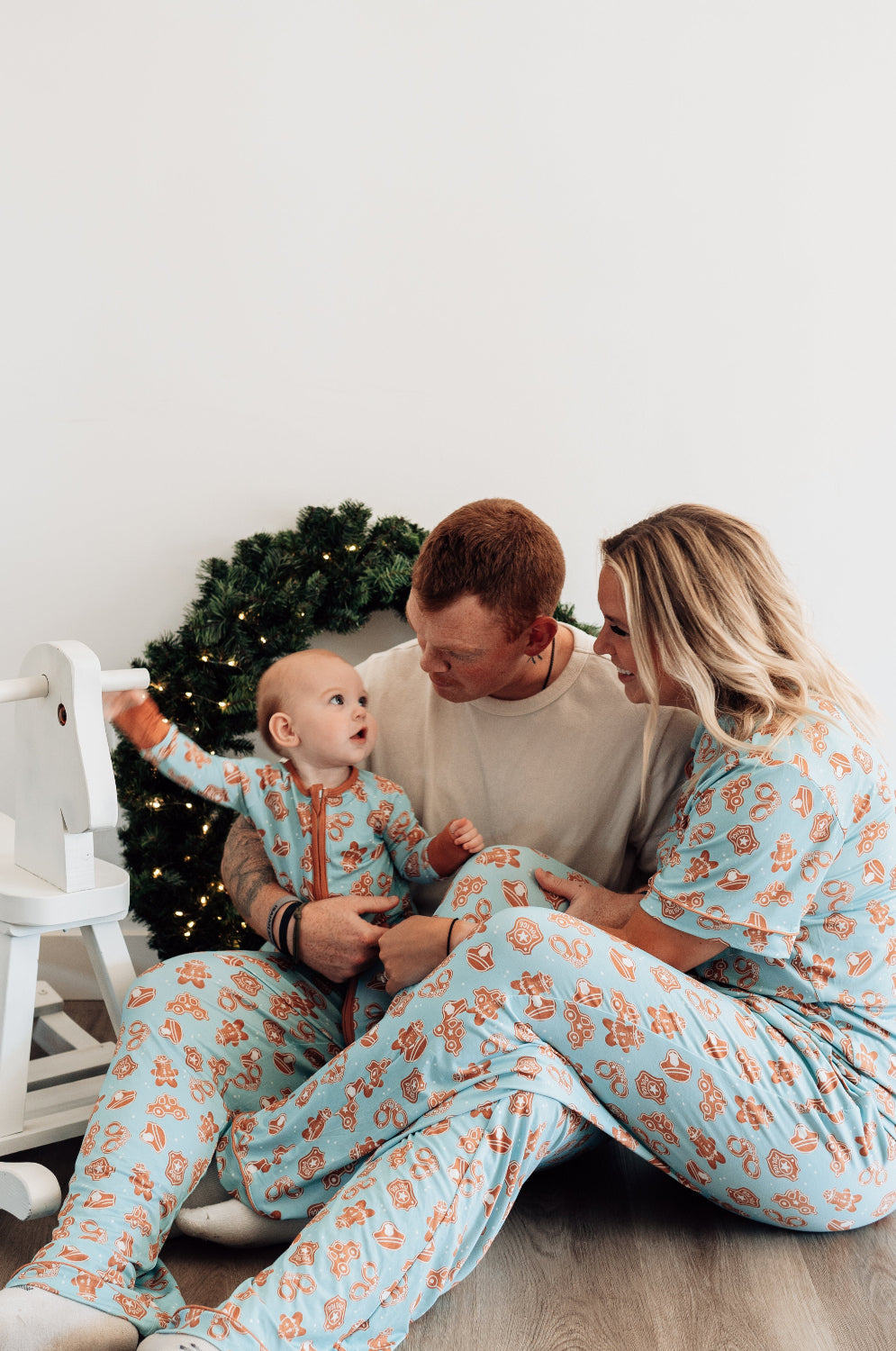 Family of three wearing matching pajamas sitting on a wooden floor with a white wall and decorative elements in the background.