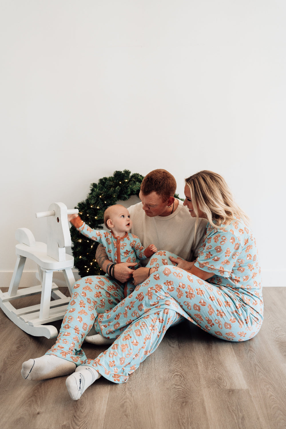 Family of three wearing matching pajamas sitting on a wooden floor with a white wall and decorative elements in the background.