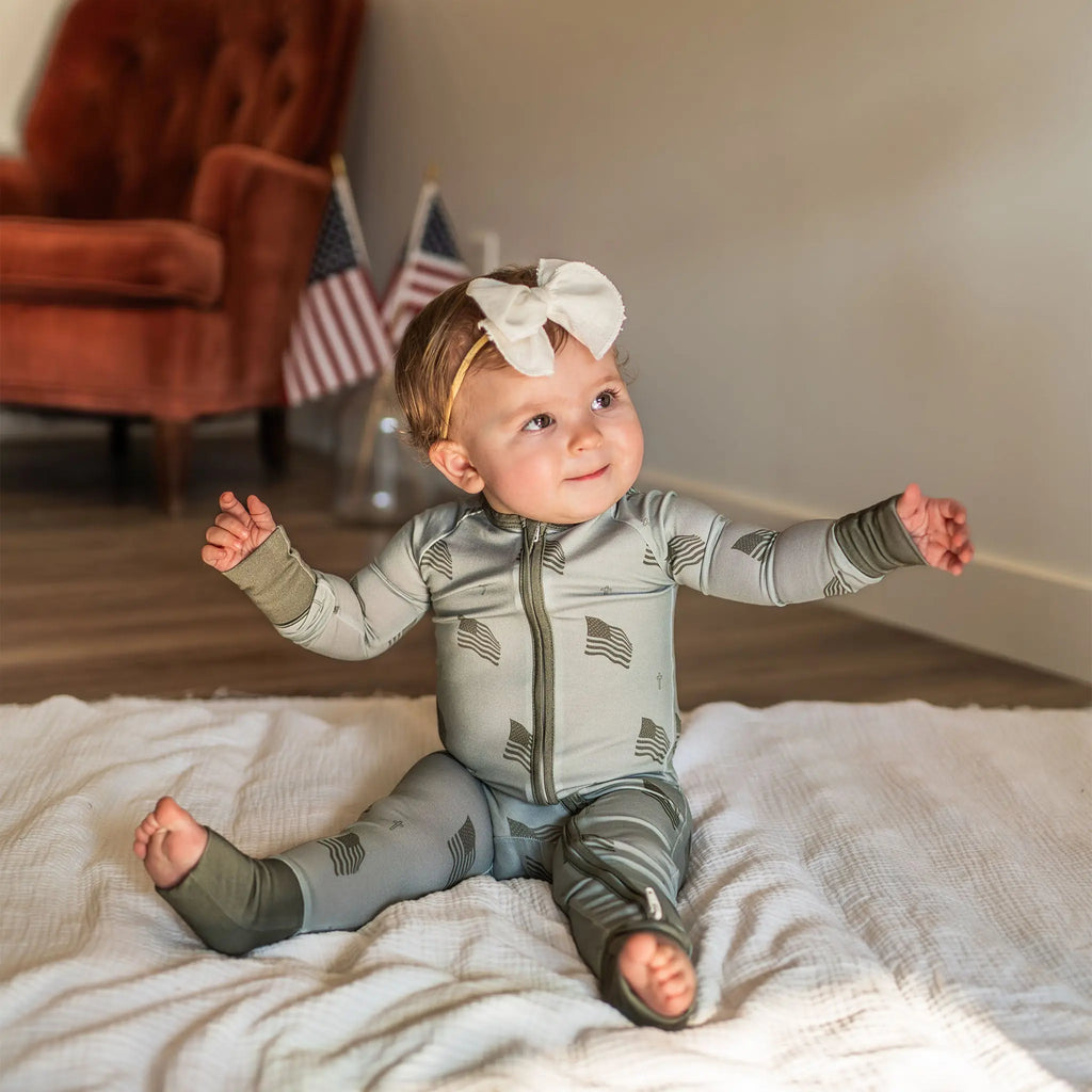 Baby sitting on a blanket wearing a patterned onesie with American flags, with a blurred background.