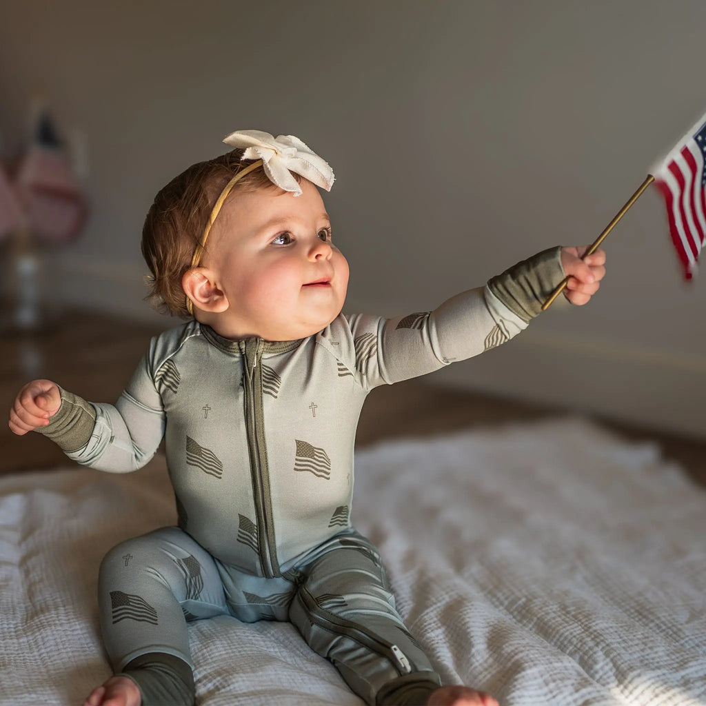 Baby in a onesie with flag pattern, holding an American flag, sitting on a bed.