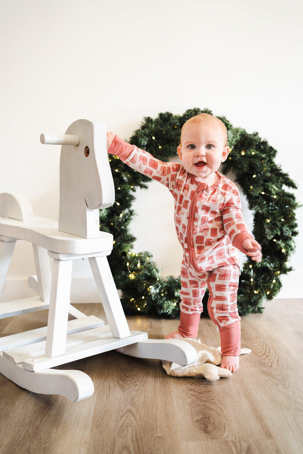 Baby in pajamas standing next to a white rocking horse with a Christmas wreath in the background.