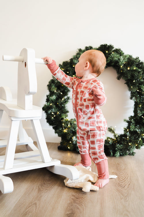 Baby in pink pajamas standing next to a white wooden toy with a Christmas wreath in the background.