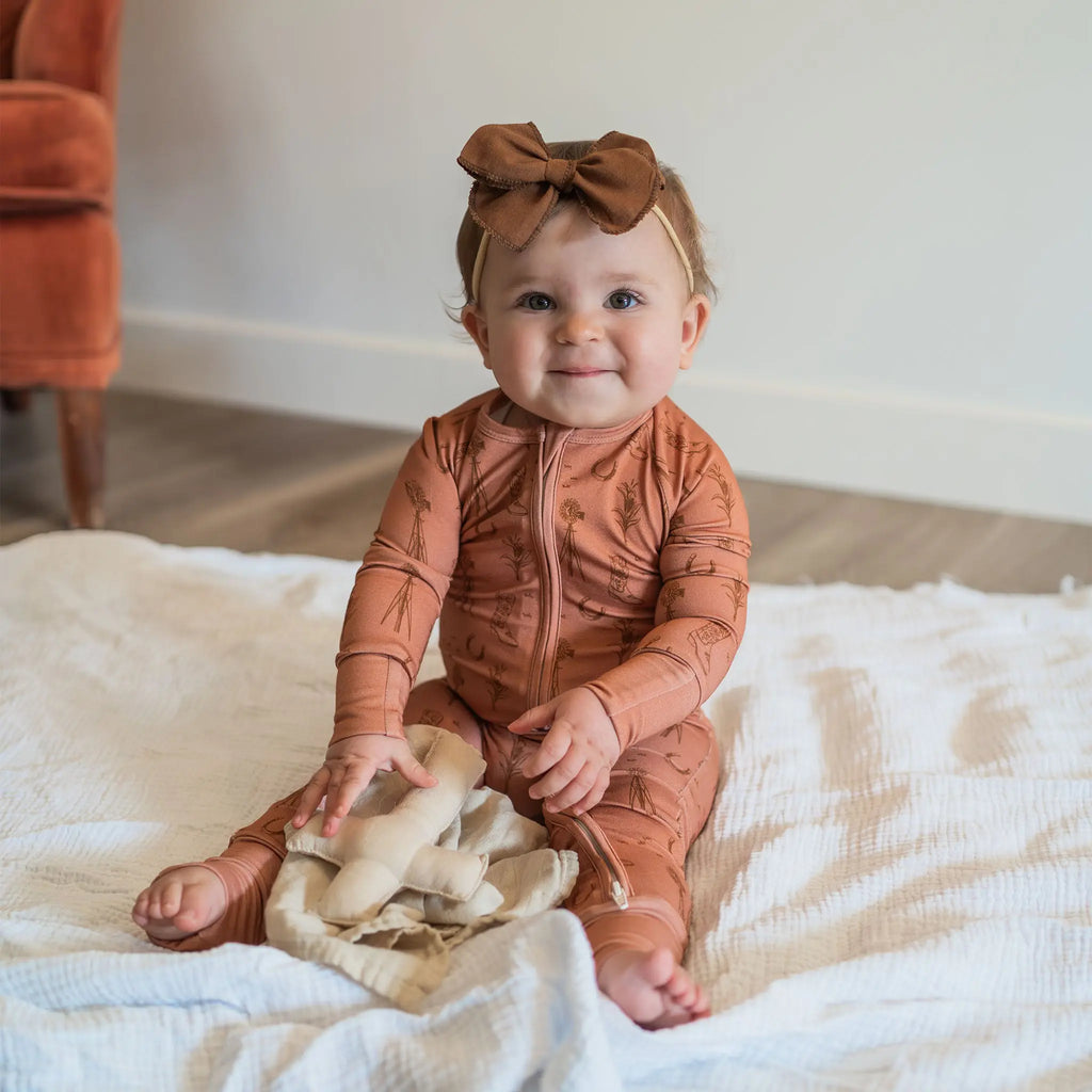 Baby in a rust-colored outfit with a bow sitting on a white blanket.