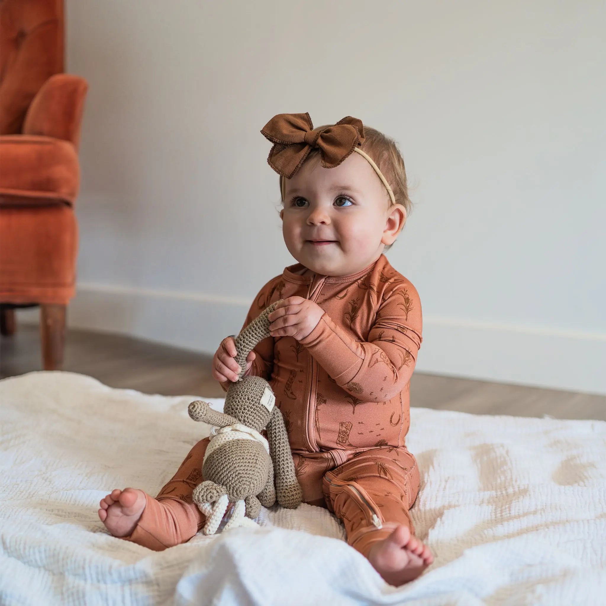 Baby in a rust-colored outfit with a bow sitting on a white blanket holding a toy.