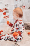 Baby wearing a onesie with red heart designs, sitting on a floor with toys around.