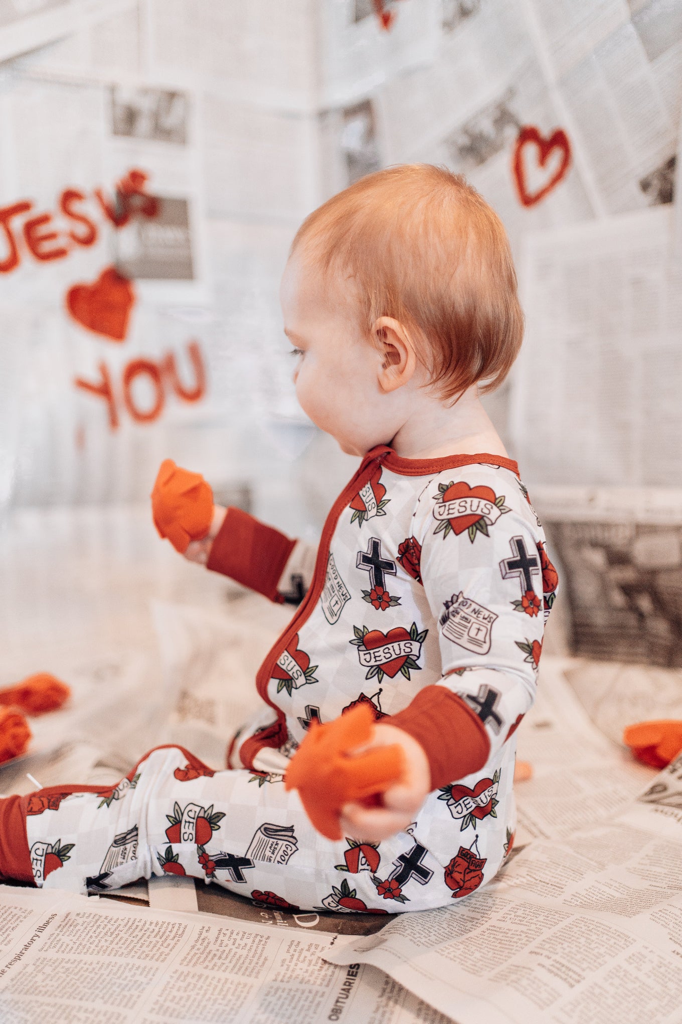 Baby wearing a onesie with red heart designs, sitting on a floor with toys around.