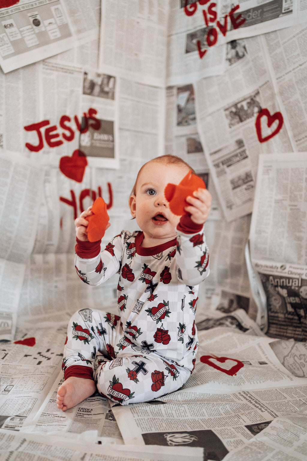 Child in pajamas holding heart-shaped objects on a newspaper background with 'Jesus Loves You' text.
