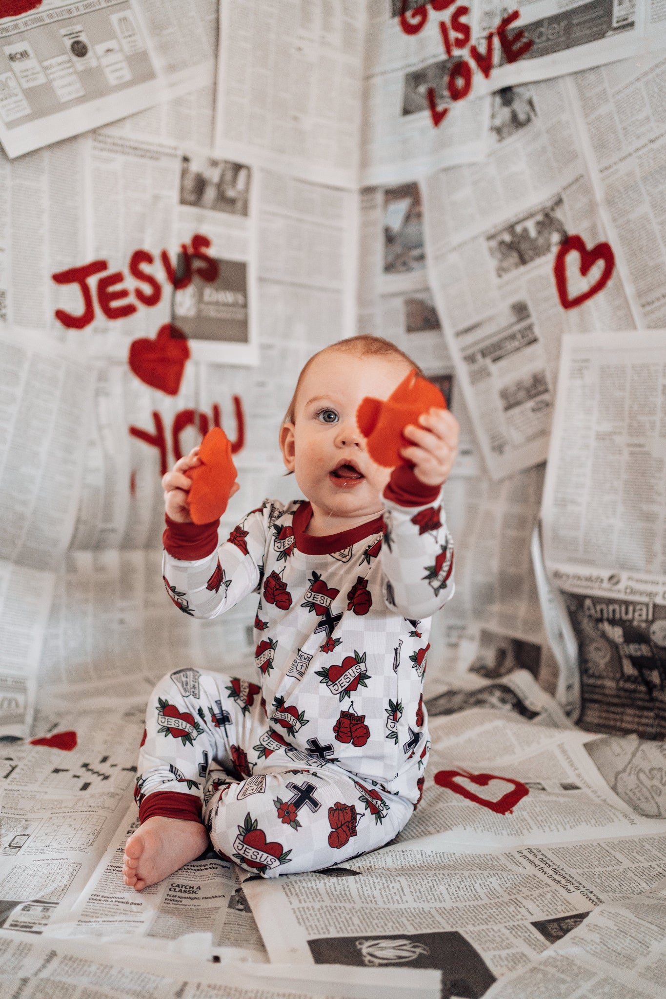 Child in pajamas holding heart-shaped objects on a newspaper background with 'Jesus Loves You' text.
