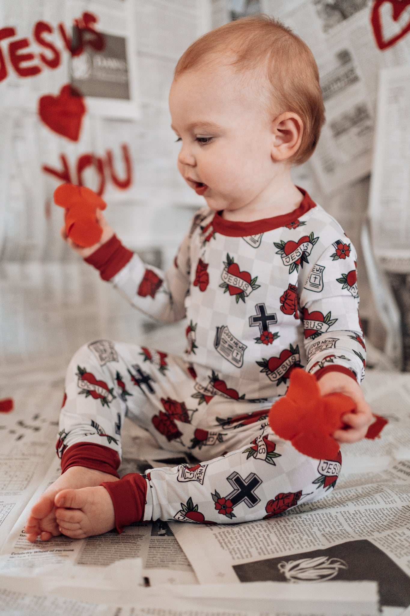 Baby wearing a pajama set with red and black designs, sitting on a bed.