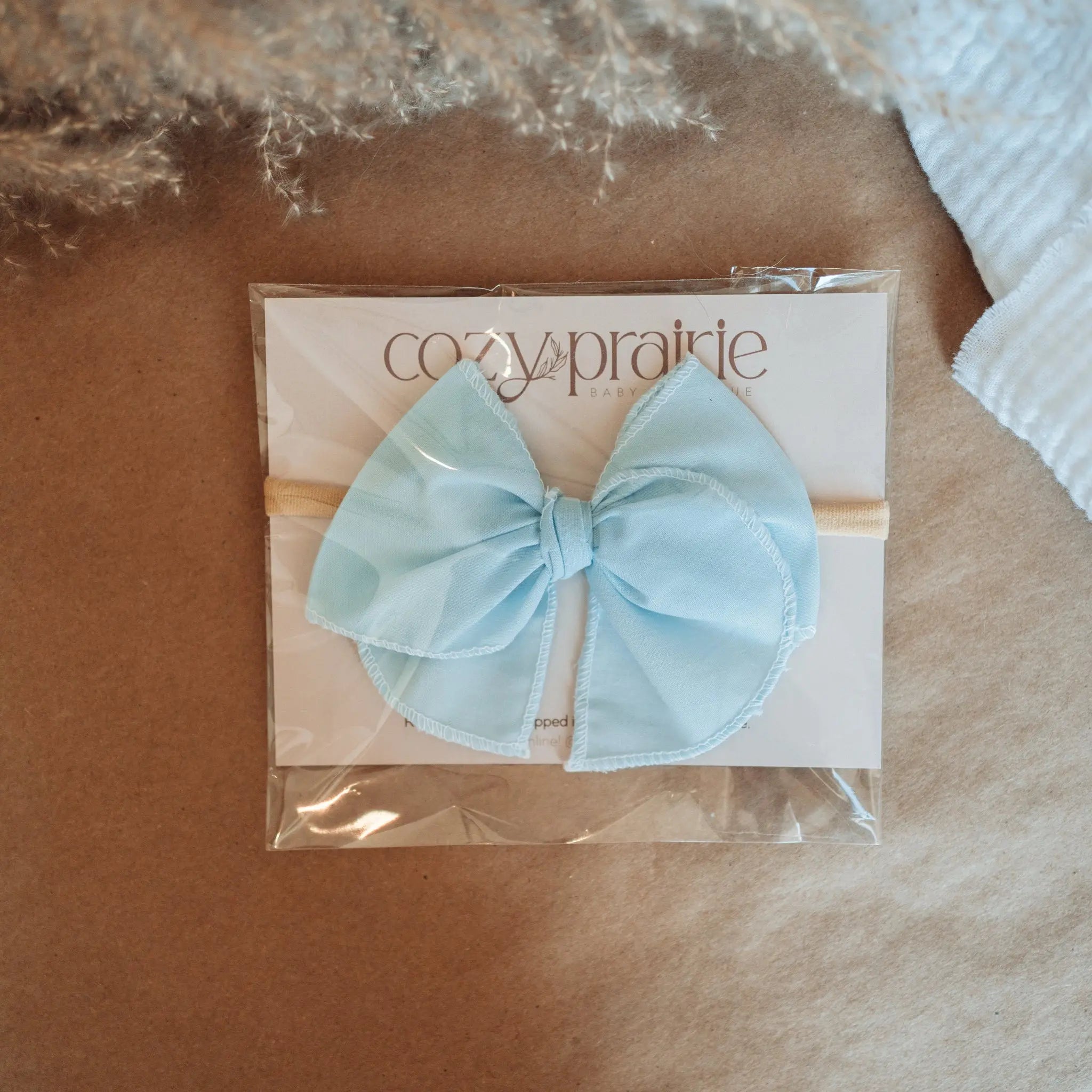 Light blue bow in clear packaging on a brown surface with 'cozy aprille' branding.