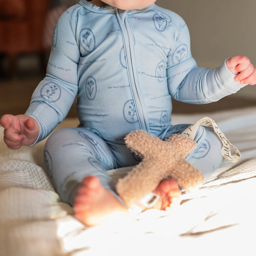 Baby in a blue onesie with teddy bear pattern sitting on a bed holding a stuffed cross.