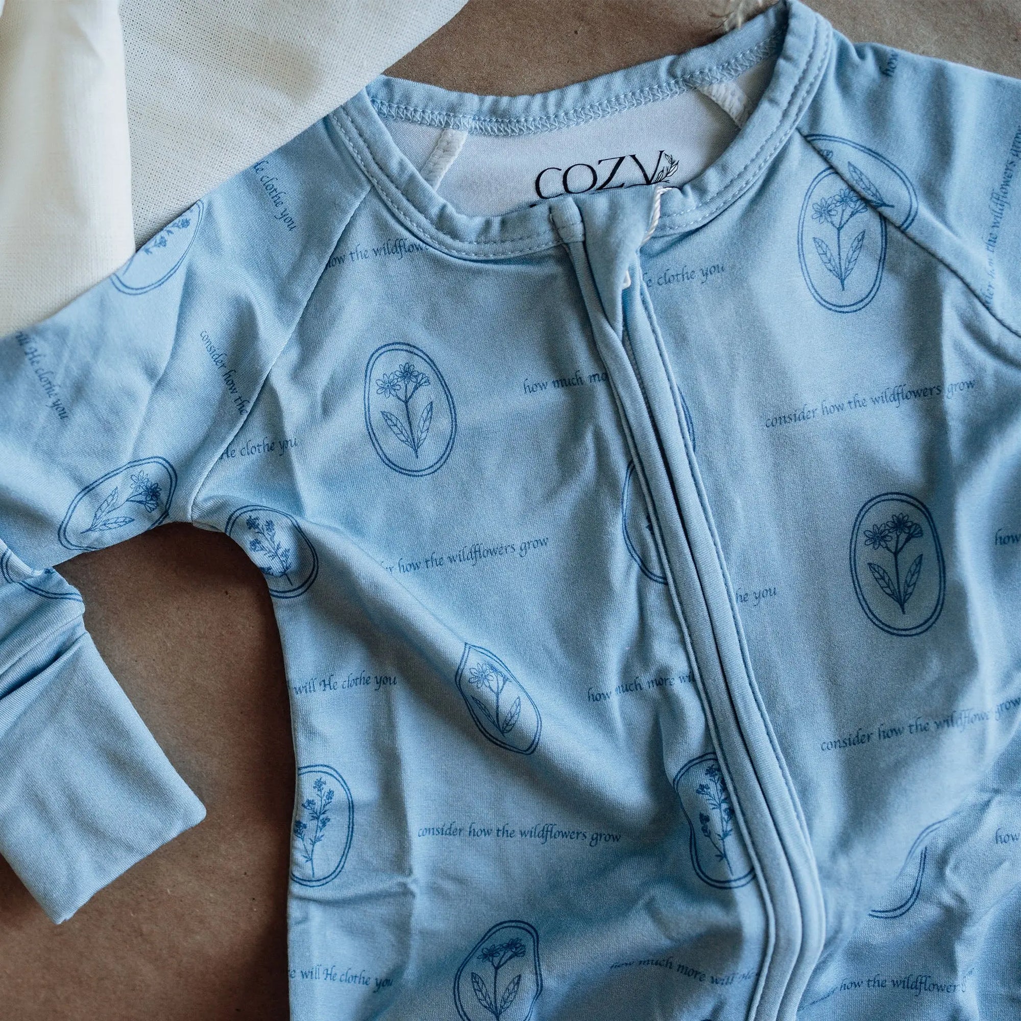 Light blue baby onesie with floral patterns and 'Cozy' branding on a wooden surface.